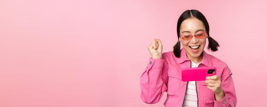 Happy smiling korean girl winning on mobile phone, looking at horizontal smartphone screen and rejoicing, achieve goal, celebrating, standing over pink background