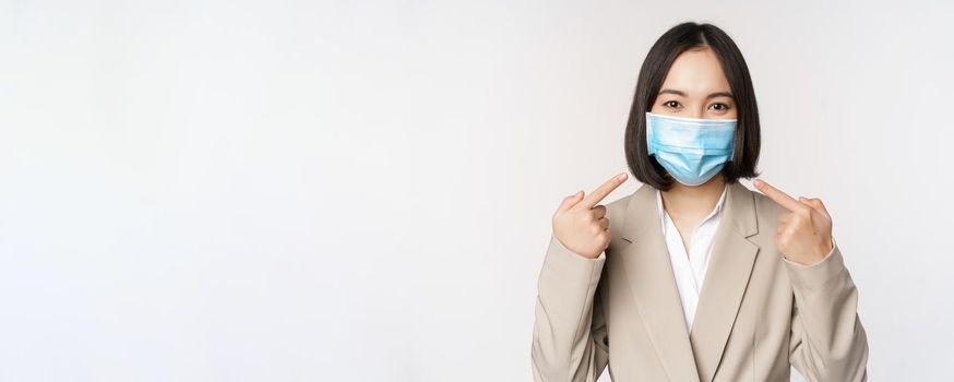 Coronavirus and business people concept. Asian female entrepreneur pointing fingers at medical face mask at workplace, standing over white background