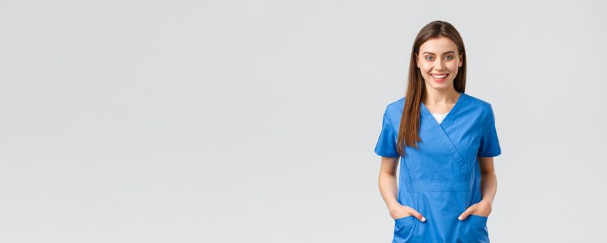 Healthcare workers, prevent virus, insurance and medicine concept. Cheerful smiling beautiful doctor, female nurse in blue scrubs, intern in hospital staying positive, standing grey background.