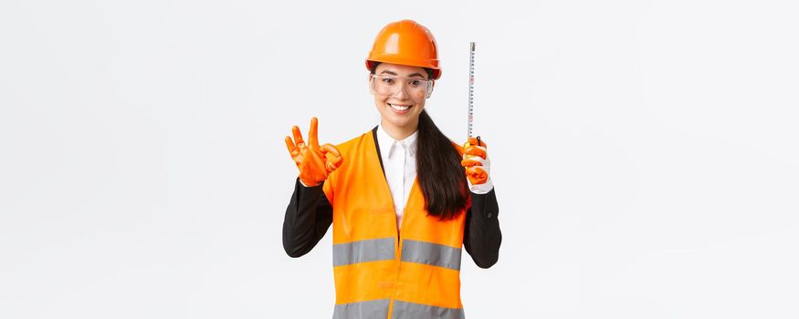 Satisfied asian female construction engineer, architect or inspector at enterprise showing okay gesture and tape measure, smiling pleased, give permission, approve measurements are valid