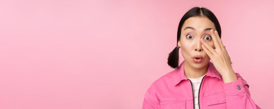Close up portrait of young asian girl looking surprised, express amazement and wonder, peeking through fingers, standing over pink background