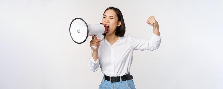 Enthusiastic asian woman, girl activist shouting at protest, using megaphone, looking confident, talking in loudspeaker, protesting, standing over white background