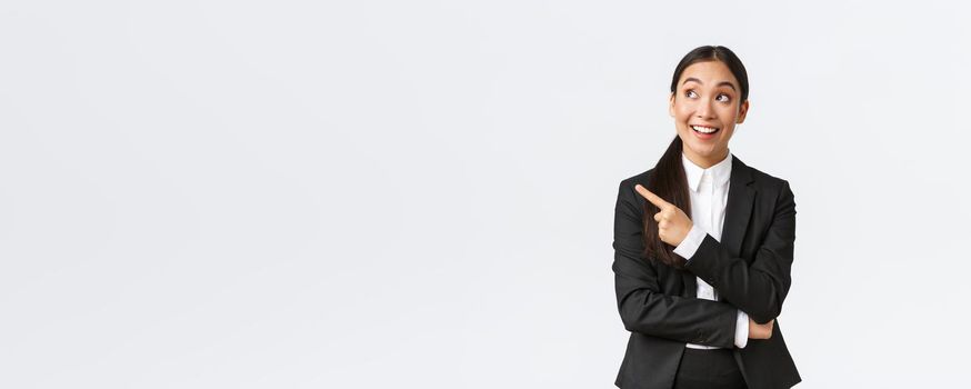 Excited and amused young asian businesswoman in suit pointing finger left and looking thrilled at promo announcement, standing white background. Entrepreneur making great suggestion