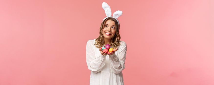 Holidays, spring and party concept. Portrait of cute and tender, lovely blond woman celebrating Easter day look dreamy up with pleased smile, holding easter painted eggs, wear bunny ears