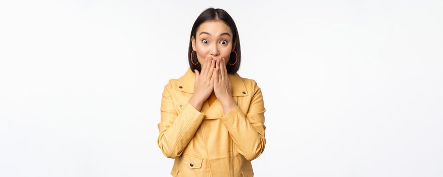 Portrait of excited asian girl looking with interest at camera, amazed reaction at big news or announcement, standing in yellow jacket over white background