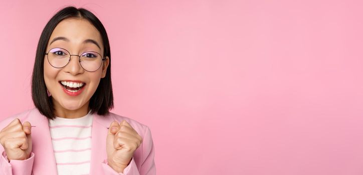 Close up portrait of asian businesswoman cheering, rooting for, looking with hope and excitement at camera, smiling and laughing, standing over pink background