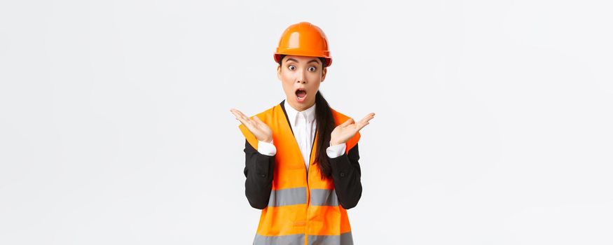 Surprised and shocked asian female engineer in reflective jacket, safety helmet, react to unexpected news, raising hands up and clap impressed, visit construction area, pleased with great work