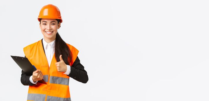 Satisfied asian female construction engineer pleased with results of inspection, wearing safety clothing and helmet at building area, writing down notes and show thumbs-up in approval