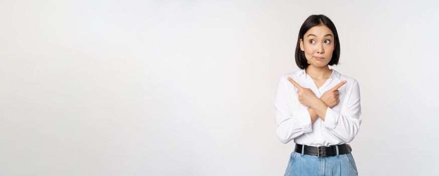 Indecisive asian woman pointing fingers sideways, pointing fingers and looking clueless, confused with choices, standing over white background