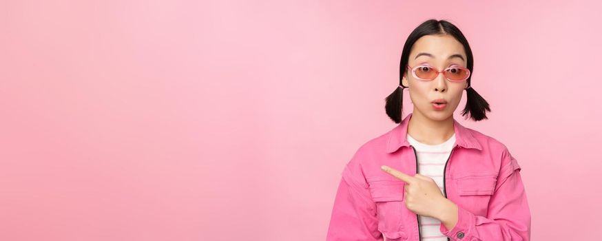 Close up portrait of asian woman gasping, looking surprised, pointing finger at banner, advertisement, standing over pink background.