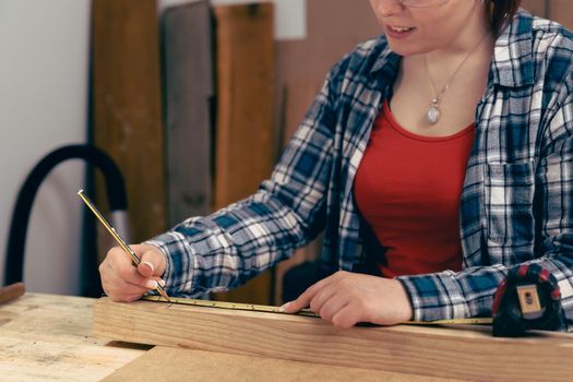 Young woman carpenter measuring a wooden board with a tape measure in her small carpentry workshop.