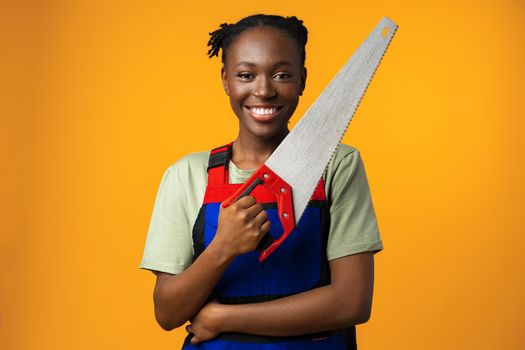 Portrait of a cute african american woman wearing uniform posing with a wood saw in her hands against yellow background