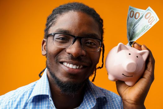 Young black african american man holding a piggy bank against orange background.