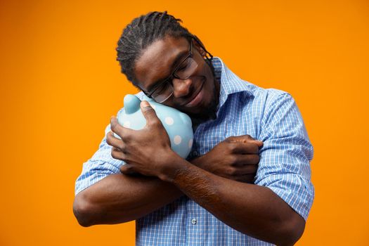 Young black african american man holding a piggy bank against orange background.
