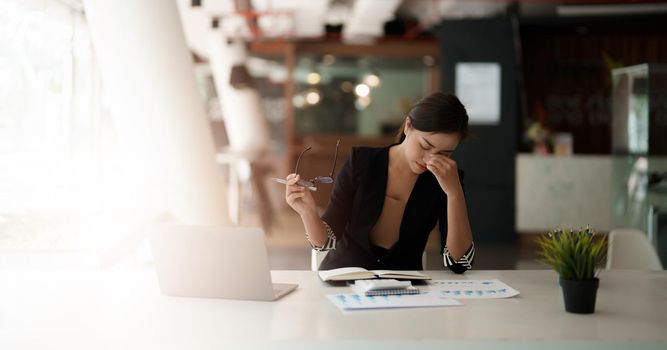 Very stressed business asian woman sitting in front of her computer looking at a large pile of paperwork, while holding a hand at her forehead