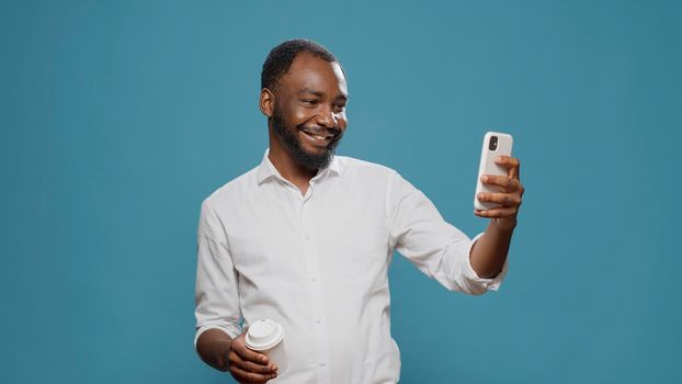 Cheerful businessman taking pictures on mobile phone, posing with coffee cup on camera. Happy freelancer using smartphone to take photos on coffee break, enjoying modern technology.