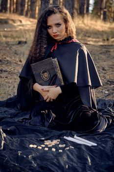 Witch in black dress with cape and hood. Posing in pine forest. Sitting on dark blanket whith books, candles and runes on it. Full length.