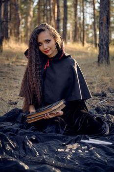 Witch in black dress with cape and hood. Posing in pine forest. Sitting on dark blanket whith books, candles and runes on it. Full length.