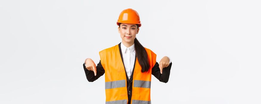 Building, construction and industrial concept. Successful female engineer, architect showing her estate project, pointing fingers down, selling houses, wearing safety helmet and reflective clothing