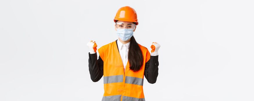 Covid-19 safety protocol at enterpise, construction and preventing virus concept. Successful winning female asian engineer in face mask, helmet and glasses triumphing, saying yes and fist pump