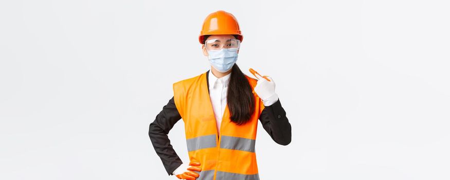 Covid-19 safety protocol at enterpise, construction and preventing virus concept. Confident asian female engineer in helmet and glasses pointing finger at face mask during coronavirus pandemic