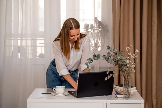 European professional woman is sitting with a laptop at a table in a home office, a positive woman is studying while working on a PC. She is wearing a beige jacket and jeans and is on the phone.