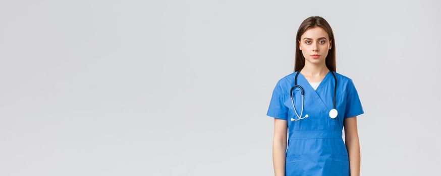 Healthcare workers, prevent virus, insurance and medicine concept. Serious-looking young nurse, doctor in blue scrubs and stethoscope, looking at camera, standing grey background