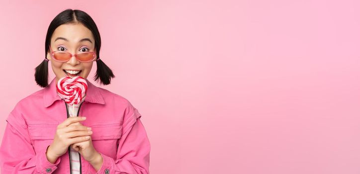 Silly and cute asian female model licking lolipop, eating candy sweet and smiling, looking excited, standing over pink background