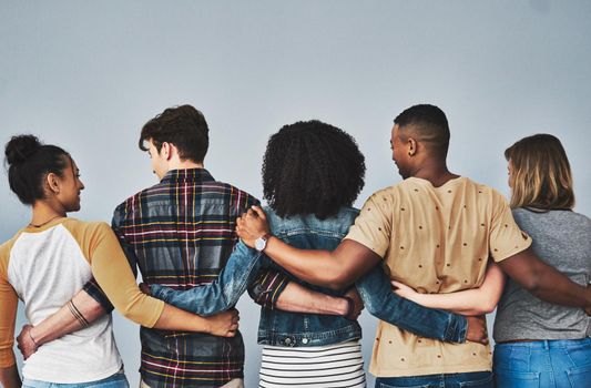 Rearview studio shot of a diverse group of young people embracing each other against a gray background.