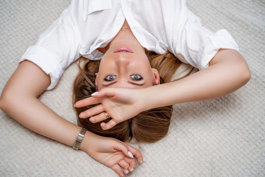 The woman lies on the bed on her back, top view. She looks straight ahead, wearing a white shirt