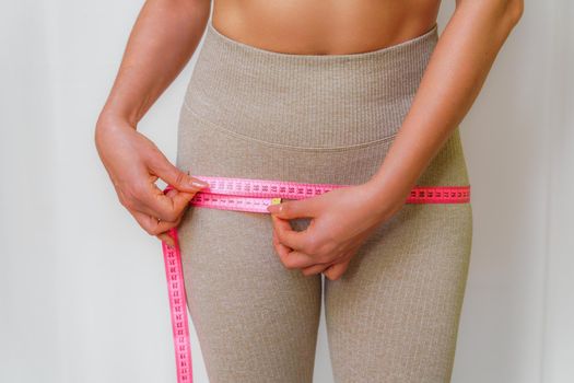 Cropped view of slim woman measuring hips with tape measure at home, close up. An unrecognizable European woman checks the result of a weight loss diet or liposuction indoors. Healthy lifestyle.