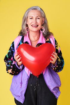 Always remember to spread love. Portrait of a confident and stylish senior woman holding a heart shaped balloon against a yellow background.