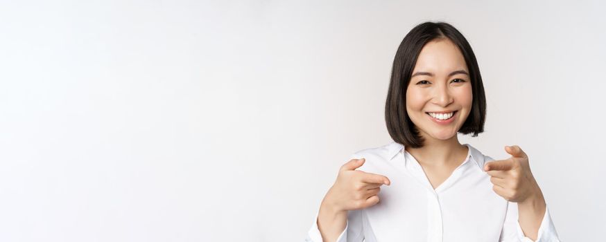Close up of enthusiastic young woman smiling, pointing fingers at camera, choosing you, inviting and recruiting people, standing over white background