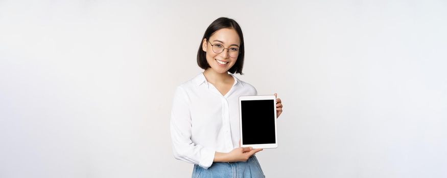 Young asian woman professional demonstrates digital tablet screen, info on her gadget, smiling and looking at camera, standing over white background