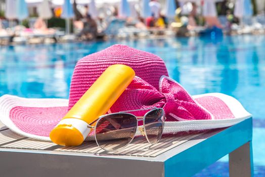 Bottle of sun cream, hat and sunglasses next to swimming pool in hotel