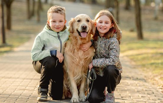 Preteen girl and boy with golden retriever dog sitting outdoors and smiling at springtime. Friends child kid with doggy pet in park