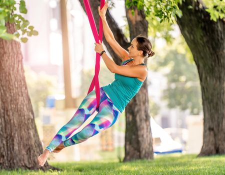 Sport girl practicing fly yoga in hammock at nature keeping her body in the air. Young woman doing aero fitness gymnastic stretching outdoors