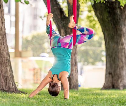 Girl during fly yoga practice in hammock stretching and relaxing her body at nature. Young woman doing aero fitness gymnastic exercising in the park