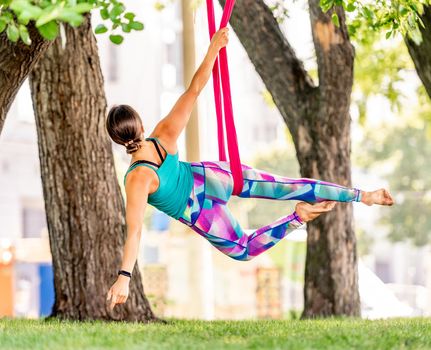 Beautiful girl during fly yoga in hammock at nature. Young sport woman doing aero fitness gymnastic stretching outdoors