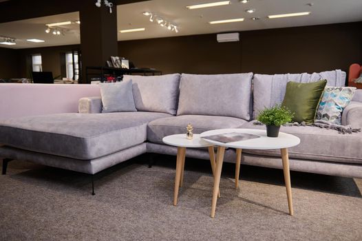 Comfortable upholstered settee with colored stylish cushions and small modern journal table displayed for sale in the furniture store showroom. Exhibition of upholstered furniture.