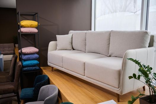 Stylish minimalist white settee with pillows displayed for sale in a showroom of modern furniture, next to stand with colorful cushions from fabrics of different texture, softness, colors and quality