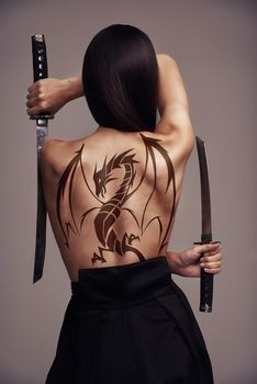 The girl with the dragon tattoo. Conceptual shot of a young female martial artist wielding two blades with a dragon tattoo on her back.