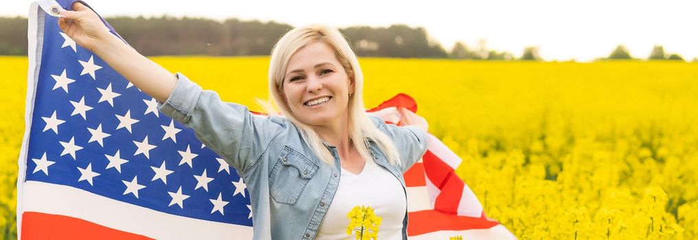 attractive woman holding an American flag in the wind in a field of rapeseed. Summer landscape against the blue sky. Horizontal orientation.