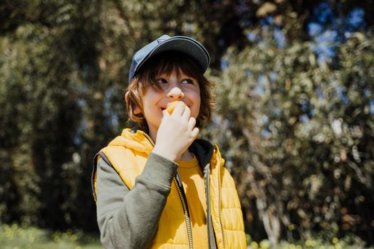 Smiling cheerful child kid in yellow vest and green hoodie eats crisp snacks outdoors in public park. Schoolboy boy enjoying consumes chews junk food outside with trees vegetation on the background