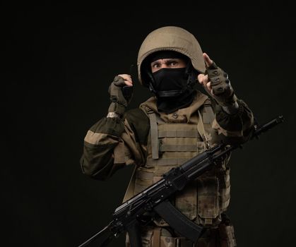a Russian soldier in military clothes with a Kalashnikov assault rifle on a dark background takes orders on a walkie-talkie