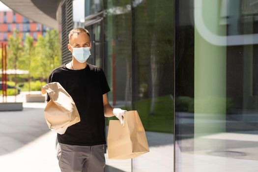 Delivery guy with protective mask and gloves holding bag with groceries in front of a building.