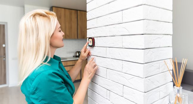 woman regulating heating temperature with a modern wireless thermostat installed on the white wall at home. Smart home heating regulation concept
