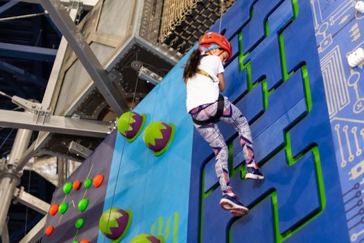 Girl wearing in harness and safety equipment climbing on practical wall indoor. Girl inserting the rope in a quickdraw.