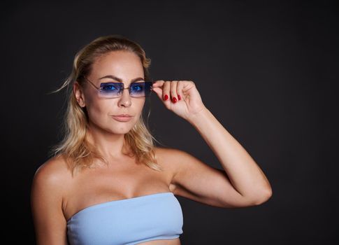 Confident portrait of a charming middle aged European woman with wavy blond hair wearing a blue top and blue sunglasses and looking at camera isolated over black background with copy ad space.
