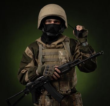 a Russian soldier in military clothes with a Kalashnikov assault rifle on a dark background takes orders on a walkie-talkie